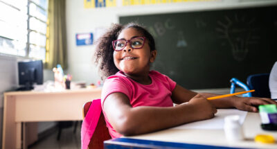 A young student with glasses thinking about gratitude and smiling at a desk in a classroom.