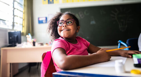A young student with glasses thinking about gratitude and smiling at a desk in a classroom.
