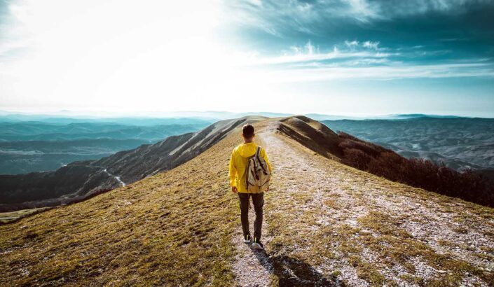 person in a yellow coat stands on an open path with water on either side and a bright sky in the distance