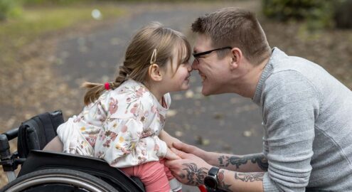 White father with glasses rubbing noses with young child sitting in a wheelchair. They look very happy.