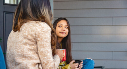 Asian teenager sitting on the porch with a parent drinking from mugs and talking.