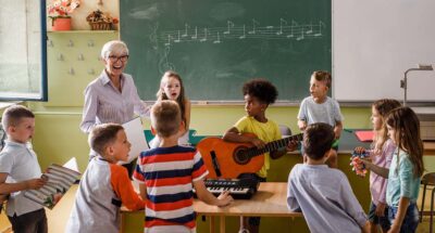 Happy senior teacher singing with large group of elementary students whole are playing musical instruments on a lass.