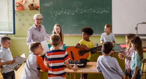 Happy senior teacher singing with large group of elementary students whole are playing musical instruments on a lass.