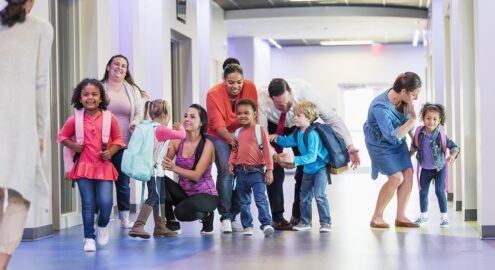 A multi-ethnic group of five preschool children, each with a parent, standing in a school hallway. The boys and girls, 4 years old, are ready for their first day of school, carrying backpacks. Their parents are giving them words of encouragement. Their teacher is walking toward them, waving.