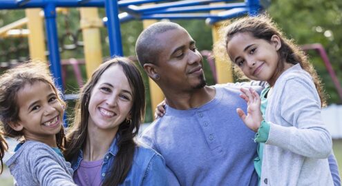A multiracial family with two girls having fun at the playground. Father is carrying the 9 year old daughter. Mother has the 7 year old. The girls are waving at the camera.