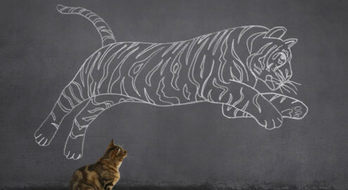 A tabby cat sitting on wooden floor and looking at the running (or jumping) tiger sketched (chalk drawing) on the wall.