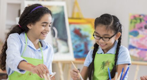 Asian and African American girls paint together during an art lesson. They are wearing aprons.