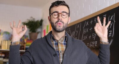 White man in glasses, flannel and sweater meditates in front of a chalkboard with math equations on it