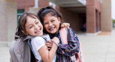 Elementary schoolgirls are playful while standing outside of the school building. they smile while playfully embracing.