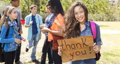 Latin descent, school girl holds Thank You sign while a multi-ethnic group of students in the background.