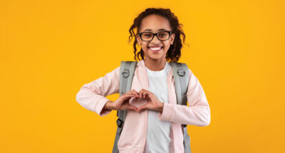 Young black girl with glasses, pink sweatshirt and backpack holds her hands up in a heart shape .