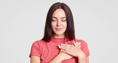 Calm brunette female with closed eyes, keeps both palms on heart, feels gratitude, being touched by something, dressed in casual pink t shirt, isolated over white background. Body language concept