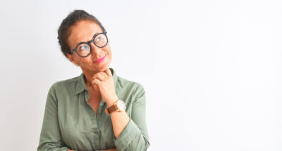 Middle age woman wearing green shirt and glasses standing over isolated white background with hand on chin thinking about question, pensive expression. Smiling with thoughtful face. Doubt concept.