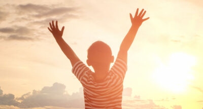 Young boy stands with arms spread in front of sunlit sky.