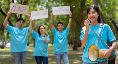 Four people in blue T-shirts outdoors, one holding a megaphone and the others holding signs that say "VOLUNTEER," "Save the World," and "NO! PLASTIC! FOAM!"