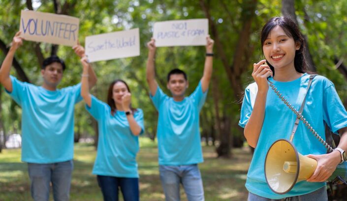 Four people in blue T-shirts outdoors, one holding a megaphone and the others holding signs that say VOLUNTEER, Save the World, and NO! PLASTIC! FOAM!