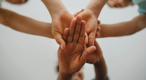 Five hands of diverse skin tones reaching together with palms touching against a light background.