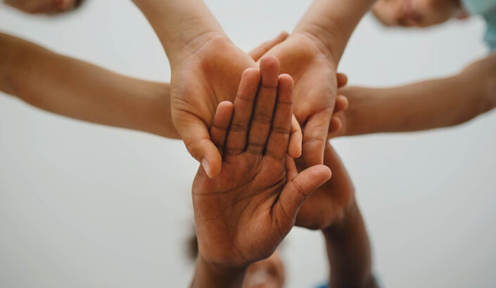 Five hands of diverse skin tones reaching together with palms touching against a light background.