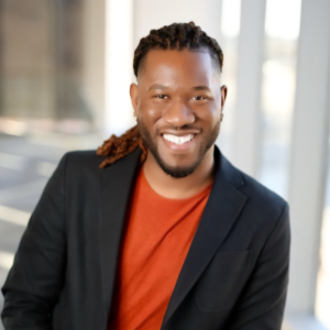 Smiling black man in his 30s looking at camera in brightly lit room, leaning forward with warm confident smile
