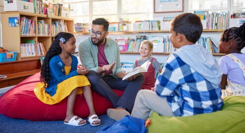 A group of children in a library, gathered around an adult holding a book, sitting on bean bags.