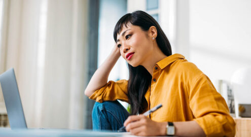 A focused woman in a casual yellow shirt working from home, using her laptop and notebook to reflect on moving forward with hope.