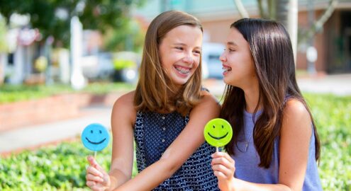 Two tweens with long hair and braces share smiley-face candies with each other.
