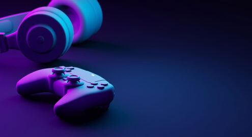 Video game controller and headphones set against a dark navy background, with purple and lighter blue lighting on them.