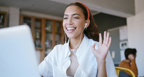 Woman with dark curly hair sits in front of a computer waving to other meeting participants