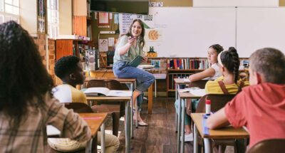 Young white teacher sits on a desk and talks through honesty scenarios with students.