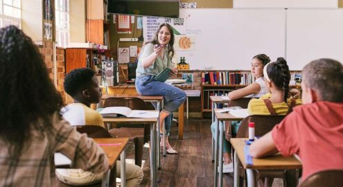 Young white teacher sits on a desk and talks through honesty scenarios with students.